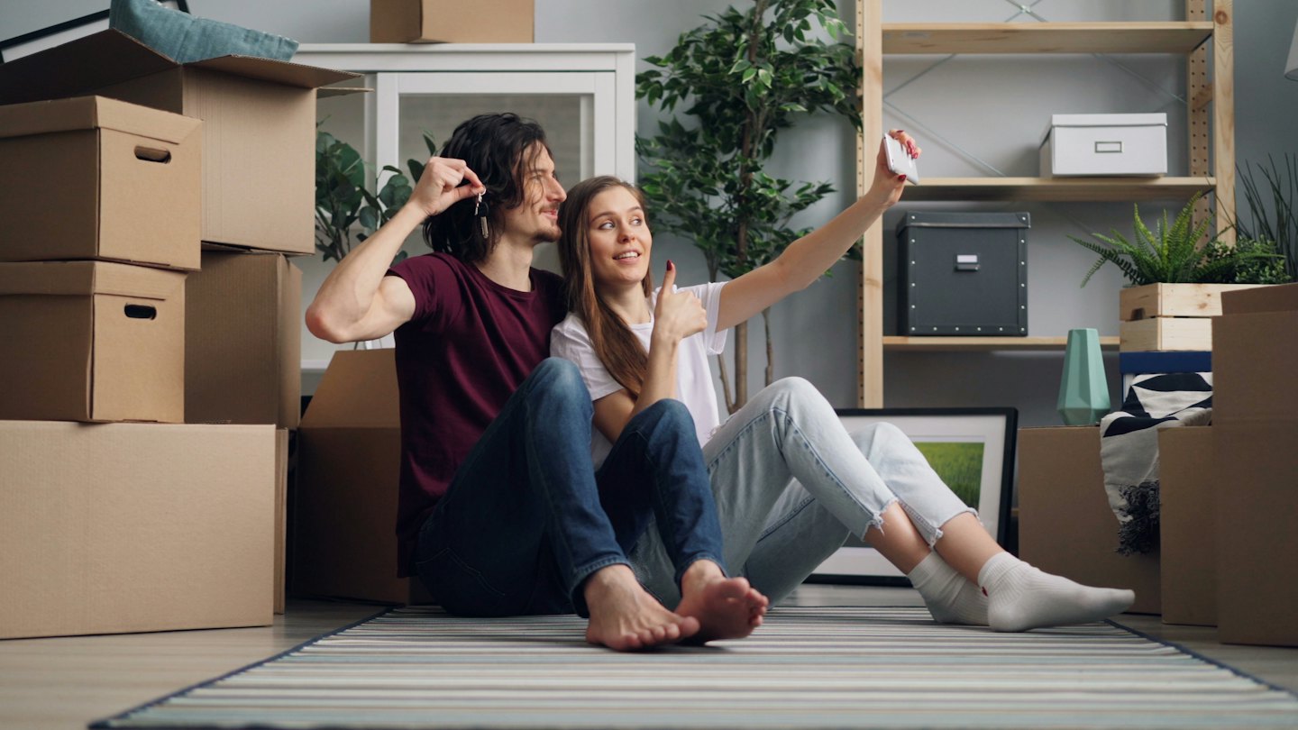 Pareja se toma una selfie con las llaves de su nuevo hogar rodeados de cajas.